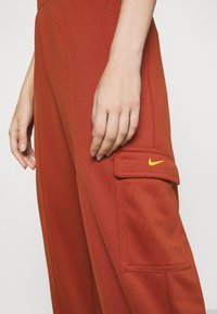 Nike cargobyxor i rostorange färg, med en sidoficka och broderad logotyp i gult. Tygmaterialet är mjukt med en svag glans.