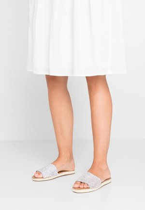 Nackte Beine, die weiße offene Slide-Sandalen mit strukturierten Riemen tragen, kombiniert mit einem weißen knieumspielenden, luftigen Rock.