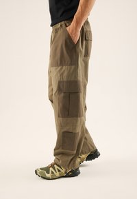 Pantalones cargo color caqui con cinturilla elástica, múltiples bolsillos de parche en diferentes tonos y un corte relajado, combinados con zapatillas deportivas verdes y beige.