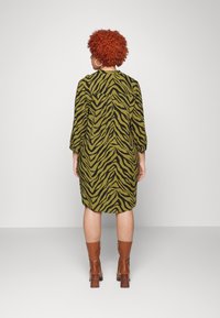 Sukienka z długim rękawem w oliwkowym kolorze z czarnym wzorem zebry, o luźnym kroju i zaokrąglonym hemie, zestawiona z brązowymi botkami na obcasie.