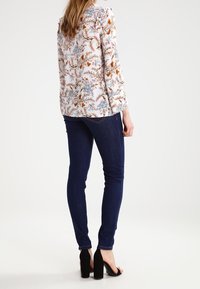 Blumenbluse mit Rüschenschultern, die auf einem hellen Hintergrund mehrfarbige Muster aufweist, kombiniert mit dunklen Skinny-Jeans und schwarzen High Heels.