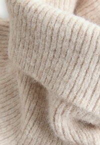 Tessuto lavorato a maglia beige con texture a coste. Il materiale appare morbido e soffice con una leggera lucentezza, mostrando diverse sfumature di crema.
