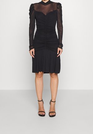 Kobieta w czarnej sukience do kolan z prześwitującymi długimi rękawami i marszczonymi detalami, w zestawieniu z czarnymi szpilkami na pasku wokół kostki, stojąca przy białej ścianie.