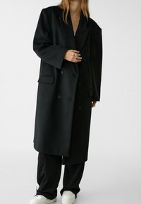 Abrigo de doble botonadura de mezcla de lana negra con corte oversized, solapas anchas, dos bolsillos delanteros y botones negros mate. Combinado con pantalones negros y zapatillas blancas.