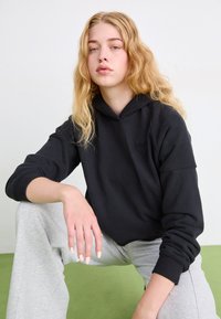 Sudadera con capucha negra de algodón, con puños de canalé. Combinada con pantalones de chándal color gris claro, ambos exhibidos sobre un fondo neutro.