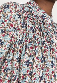 Camicia a motivi floreali in blu chiaro, crema e rosso, caratterizzata da pieghe e dettagli con bottoni, realizzata in un tessuto leggero.