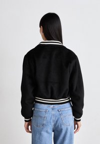 Sort jakke med ribbet hvidstribet krave og manchetter, blød tekstur, cropped pasform, parret med lyseblå højtaljede jeans.