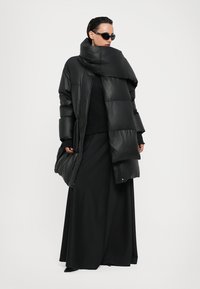 Μαύρο φουσκωτό παλτό με ψηλό γιακά, με γυαλιστερή φινίρισμα και υπερμεγέθη σιλουέτα, σε συνδυασμό με μια μακριά μαύρη φούστα.