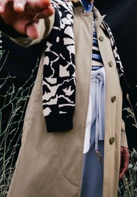 Beige trensscoat, jossa on suuret napit, yhdistetty musta-kermanväriseen kuvioituun neuleeseen. Kerrostettuna vaaleansiniselle vaatteelle, jossa on solmiyksityiskohtia.