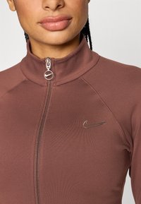 Καφέ ζακέτα με φερμουάρ από λείο ύφασμα. Διαθέτει ψηλό γιακά, εφαρμοστή γραμμή και μικρό μεταλλικό λογότυπο της Nike στο αριστερό στήθος.