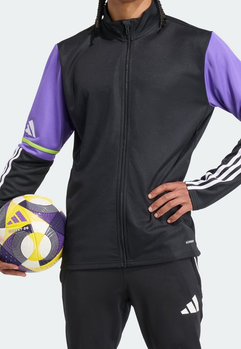 Personne portant une veste noire avec des manches violettes tenant un ballon de football Adidas violet, jaune et blanc à hauteur de taille, main sur la hanche.