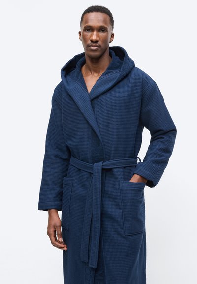 Άνδρας που φοράει μπλε ναυτικό μπουρνούζι με υφή, κουκούλα και ζώνη, με το ένα χέρι στην τσέπη, στέκεται μπροστά σε απλό φωτεινό φόντο.