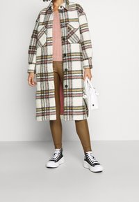 Kraciasty wełniany płaszcz w kolorach białym, beżowym i różowym; długi fason z zapięciami na guziki i kieszeniami. Do tego brązowe leginsy i czarne trampki.