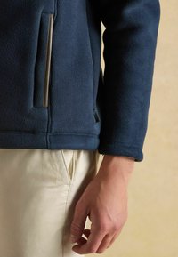 Tmavomodrá fleecová bunda so zipsom a kontrastným béžovým lemovaním; má hladkú textúru a uvoľnený strih, skombinovaná so svetlými nohavicami.