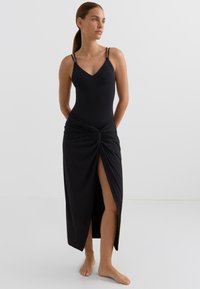 Črna obleka z oprijetim korzetom in detajlom zavezovanja v pasu ter visokim razporkom na stegno. Tanke naramnice in mehka tekstura tkanine.