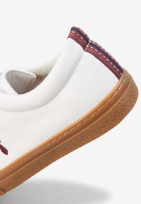 Zapatilla de piel blanca con suela de goma texturizada, con un acento de tela burdeos en el talón y costuras decorativas a lo largo del borde.