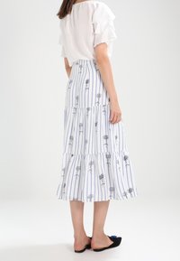 Žena v bílé off-shoulder blůze a bílé sukni s vertikálními modrými pruhy a černými květinovými vzory, doplněná černými plochými botami.