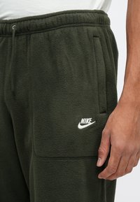 Zelené flísové tepláky s elastickým pasem, bočními kapsami a bílým logem Nike na levém stehně. Měkká textura a volný střih.