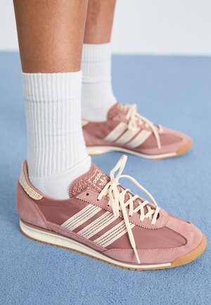 Osoba nosząca białe prążkowane skarpetki i różowe trampki Adidas z beżowymi paskami, stojąca na niebieskim wykładzinie.