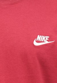 Rdeča tkanina v bližnjem posnetku z belim izvezenim napisom Nike in logotipom Swoosh na desni strani.