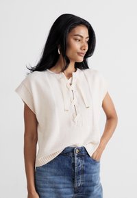 Light beige knitted top with short sleeves, lace-up front detail, and textured patterns. Paired with high-waisted blue denim jeans.