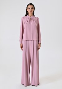 Blusa rosa a maniche lunghe con scollo plissettato abbinata a pantaloni a gamba larga, realizzati in tessuto morbido. Il design presenta una leggera lucentezza.