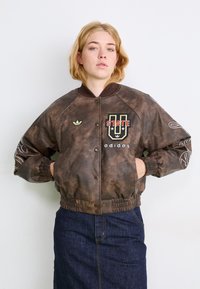Rjavo usnjeno bomber jakno iz umetnega usnja s rebrastimi zapestniki in spodnjim delom, vezenim logotipom in zapiranjem na gumb. Ujemajoča usnjena krila.