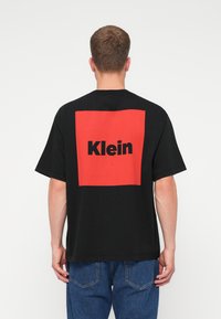 Crna pamučna majica s velikim crvenim kvadratom na leđima na kojem je riječ "Klein" ispisana masnim crnim fontom. Ležeran kroj s kratkim rukavima.
