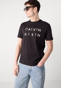 Tricou negru din bumbac cu logo-ul „CALVIN KLEIN” în stil îndrăzneț și finisaj metalic, asociat cu blugi denim de culoare bleu deschis. Modelul poartă ochelari de soare închiși la culoare.