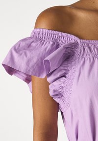 Dunkelhäutige Person in einem lilafarbenen, schulterfreien Oberteil mit gerafftem elastischem Halsausschnitt und kurzen, flatternden Ärmeln, vor einem weißen Hintergrund.