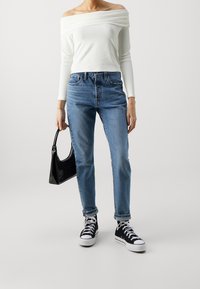 Weißes schulterfreies langärmliges Oberteil, blaue hochgeschnittene Jeans, schwarze lackierte Handtasche und schwarze High-Top-Sneaker mit weißen Sohlen.