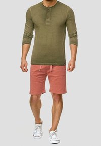 Man in een olijfgroene lange mouwen henley-shirt, koraalkleurige short met aantrekkoord en witte sneakers die tegen een plain achtergrond loopt.