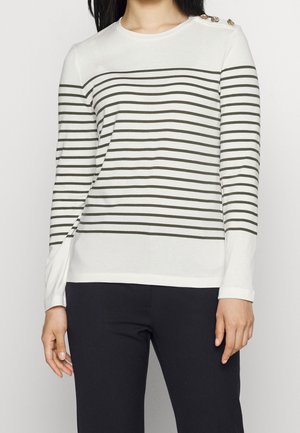 Femme portant une chemise blanche à manches longues avec des rayures horizontales noires et des boutons dorés sur l'épaule gauche, associée à un pantalon noir.