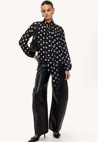 Blouse noire à pois avec un col noué, manches larges, associée à un pantalon large en cuir noir brillant et à des talons pointus. Sac à main noir.