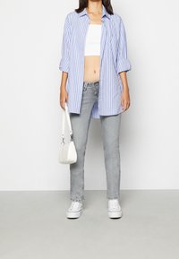 Mujer con una camiseta blanca corta, una camisa larga a rayas azules y blancas, pantalones vaqueros grises, zapatillas blancas y sosteniendo un pequeño bolso blanco.