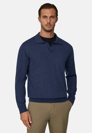 Man draagt een blauwe trui met lange mouwen over een zwart hemd en beige broek, staat met één hand in de zak.