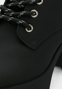 Gros plan d’une chaussure noire avec des lacets noirs et blancs à motif, passés dans des œillets argentés sur une tige en cuir lisse.