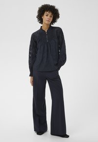 Femme debout portant un chemisier noir à manches longues texturé avec des détails plissés et un pantalon large et sombre, sur un fond uni.