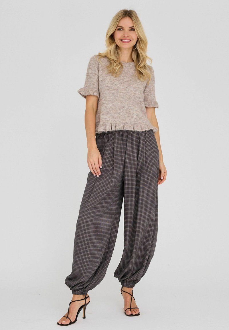 Femme souriante portant un top en maille beige à manches courtes et un pantalon large à carreaux foncés avec des poignets élastiques et des talons hauts noirs à brides.