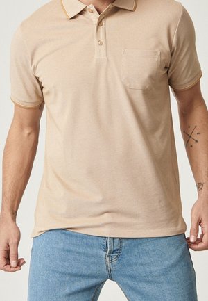 Uomo che indossa una polo beige a maniche corte con tasca sul petto e jeans azzurri, in piedi con le braccia leggermente piegate.