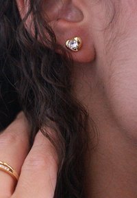 Orecchini a forma di cuore in oro con una pietra preziosa trasparente. Superficie liscia con una finitura lucida, posizionati vicino a un ciuffo di capelli ricci.