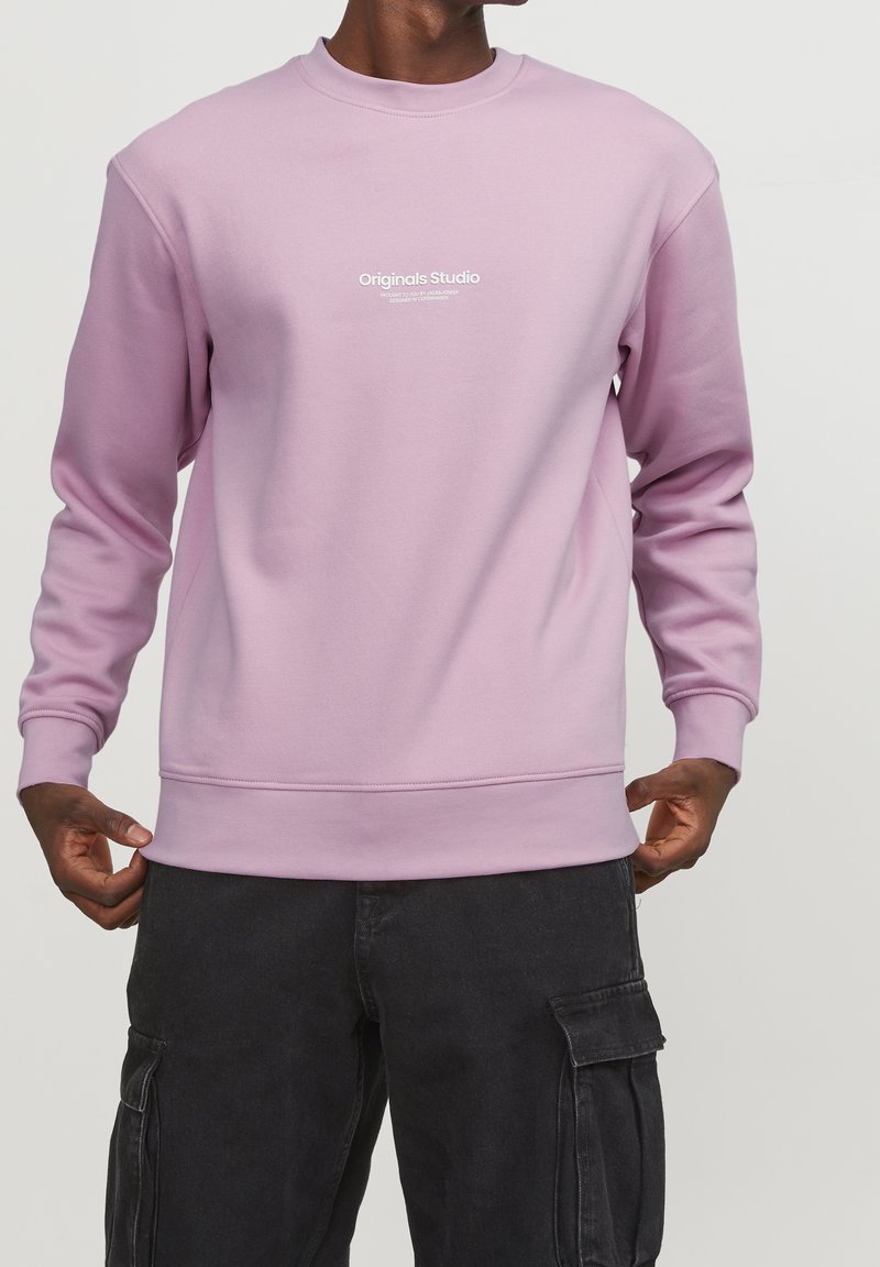 Sudadera rosa claro con cuello redondo, puños y cintura acanalados. Presenta el logo "Originals Studio" impreso en el frente. Textura suave.
