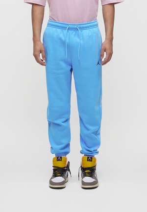 Uomo che indossa pantaloni della tuta Jordan azzurri e sneakers Air Jordan multicolori, in piedi su sfondo bianco.