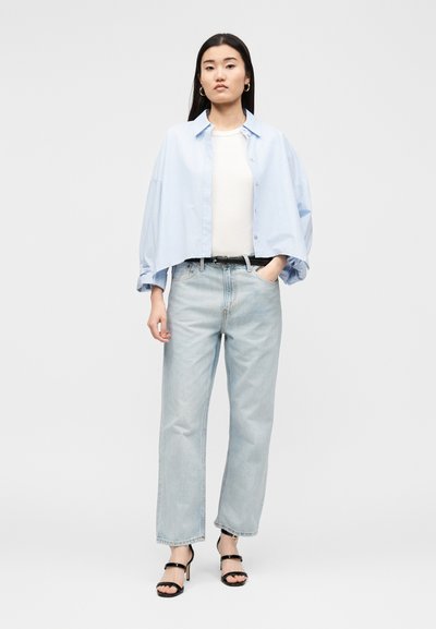 Γυναίκα στέκεται φορώντας ένα ανοιχτό μπλε φαρδύ πουκάμισο, λευκό τοπ, ανοιχτό μπλε τζιν, μαύρη ζώνη και μαύρα σανδάλια με τακούνι σε απλό φόντο.