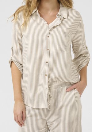 Mujer que lleva una camisa de botones de rayas verticales beige y blancas con las mangas remangadas y pantalones a rayas a juego con la mano en el bolsillo.