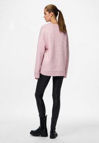 Rožnati pleteni pulover z ohlapnim krojem, rebrastim robom in spuščenimi rameni, kombiniran s črnimi legicami iz umetnega usnja in krepko črnimi škornji.