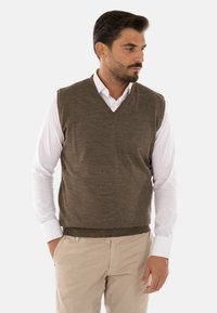 Maglione senza maniche in lana marrone con scollatura a V, indossato sopra una camicia bianca con bottoni, abbinato a pantaloni beige. Texture liscia.
