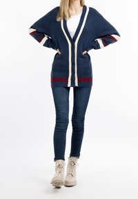 Marinblå cardigan med krämfärgad kant och röda ränder, med knäppning fram och oversized design. Kombinerad med mörka jeans och beiga snörstövlar.