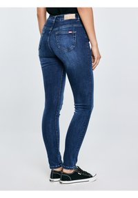 Sieviete, kas valkā tumši zilas šauras džinsu bikses un melnus zema profila sporta apavus, stāv pret vienkāršu gaišu fonu.