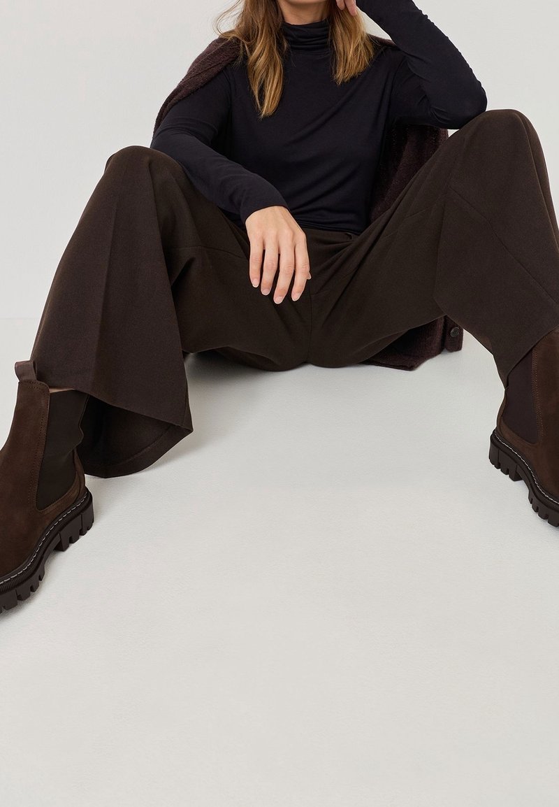 Femme assise par terre, jambes écartées, portant un pantalon large marron foncé, des bottes assorties, un col roulé noir, et un manteau sombre posé sur les épaules.